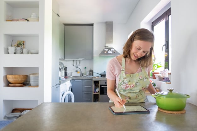 mujer joven positiva escribiendo notas receta pantalla pad usando tableta cerca cacerola grande mostrador vista frontal espacio copia cocinar casa e internet concepto 74855 7761